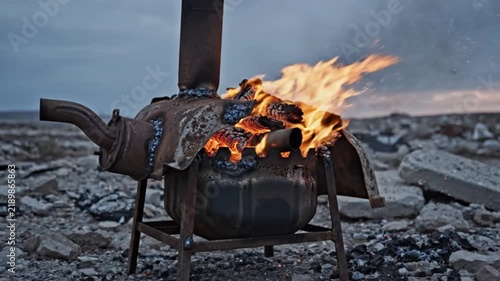 Rustic makeshift metal stove burning brightly outdoors in a rocky landscape during a cold overcast day