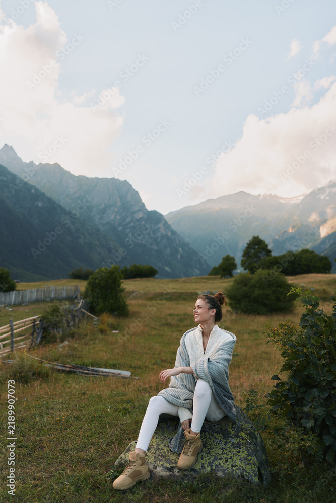 Obraz premium woman enjoying nature in mountainous meadow, sits on rock, wearing cozy cardigan and boots, peaceful outdoor lifestyle portrait with scenic landscape and natural light