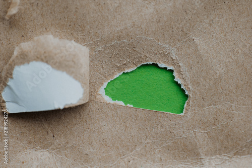 Fotografie Small torn hole in kraft paper revealing green background for teaser campaigns, hidden messages, investigations concepts