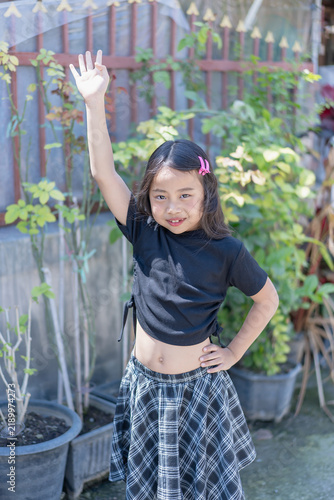 Happy young girl raising her hand outdoors in a garden, smiling at the camera in bright sunlight. Childhood, fashion, lifestyle, summer, nature, playful activity, confidence, and joyful youth concept