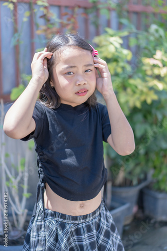 Young girl adjusting her hair outdoors in bright sunlight, wearing a black shirt and plaid skirt, with a natural portrait expression. Childhood, fashion, lifestyle, summer, and happy youth outdoors.