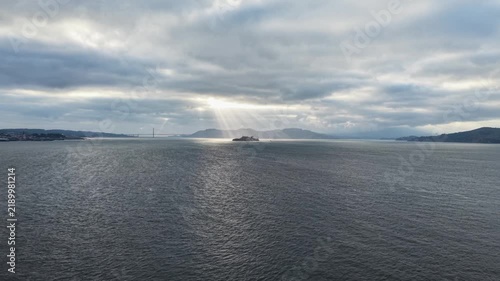 Aerial rising vertically over the San Francisco Bay, revealing Alcatraz Island illuminated by dramatic sun rays under an overcast daytime sky