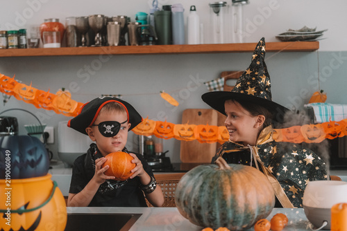 Portrait of a girl and her a little brother celebrating Halloween and decorating pumpkins in the kitchen. They look scary in witch and pirate costumes. Kids Halloween party.