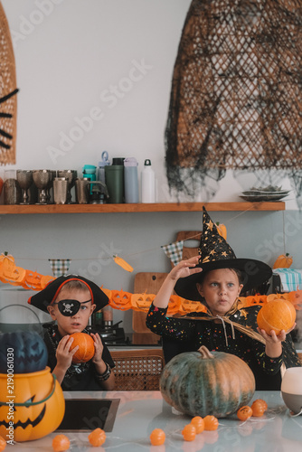 Portrait of a girl and her a little brother celebrating Halloween and decorating pumpkins in the kitchen. They look scary in witch and pirate costumes. Kids Halloween party.