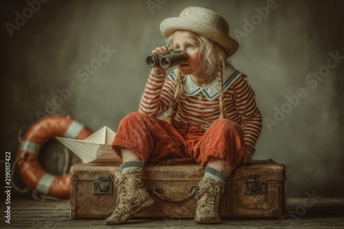 Young girl wearing a straw hat and striped shirt sits on a vintage suitcase, holding binoculars, with a paper boat and lifebuoy in the background