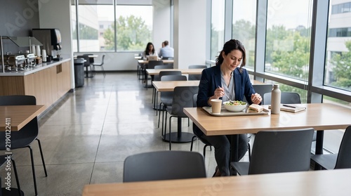 Professional businesswoman sitting alone in a modern office cafeteria eating a healthy salad and sandwich during her lunch break.