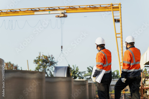 Engineers control crane lifting prefabricated metal cylinder at construction factory yard. Concept of heavy manufacturing, rigging operations, industrial logistics and engineering management.
