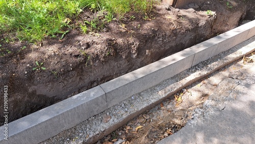 Concrete curb installation along a dirt trench with grass and soil visible, showcasing construction progress on a sidewalk or road project