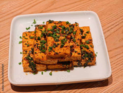 Chinese pan-fried spicy tofu topped with chopped green onions and chili flakes, served on white plate upon a wooden tabletop.