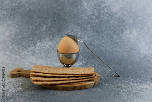 Принти на полотні Cup holds boiled egg with spoon placed next to crispbread on wooden board on gra