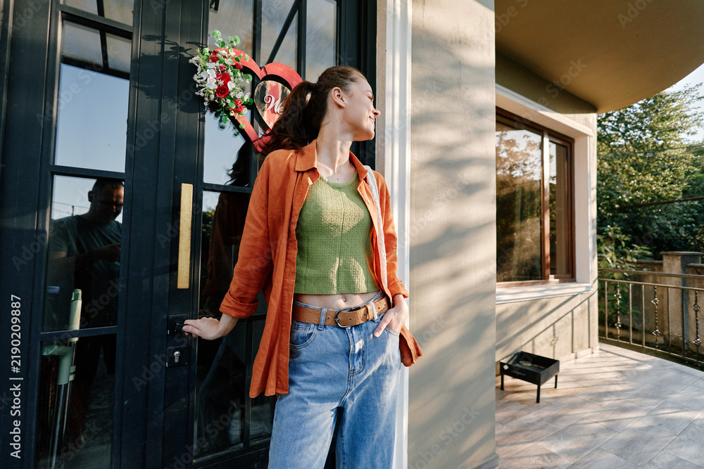 Obraz premium Young woman in orange shirt and green top standing at a black door with a heart wreath. Side view of a stylish casual outfit. Concept of fashion, spring style, and lifestyle.
