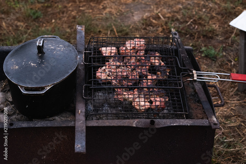 Canvastavla Juicy meat grilling in a metal wire grate over hot coals with black cast iron pot on BBQ grill outdoors