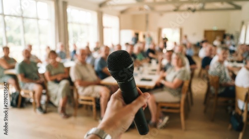 A hand holds a black microphone in front of a large group of people seated at tables in a bright, airy hall. The background shows a diverse crowd of adults listening attentively.