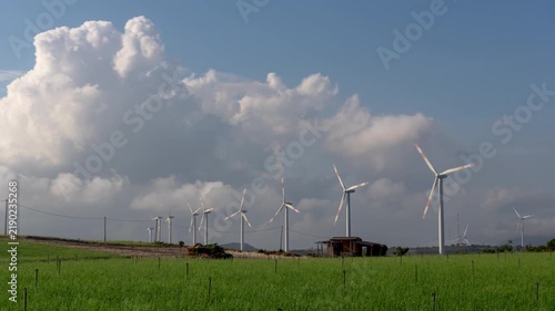 Timelapse of modern wind turbines spinning in a green field under a dynamic blue sky with fluffy white clouds.