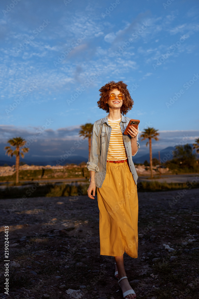 Fototapeta premium woman outdoor lifestyle portrait casual fashion sunny nature backdrop palm trees summer smiling device wind breeze color tone earthy calm leisure confidence denim jacket dress candid moment