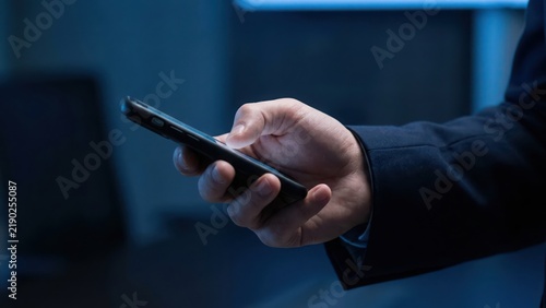 Close up of a businessman in a dark suit holding a modern smartphone in his hand, browsing digital in a dimly lit office environment setting