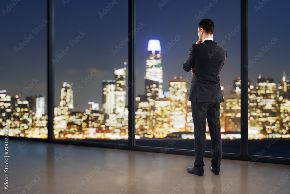 Fototapeta premium Businessman standing by a window at night, viewing a glowing city skyline while reflecting on corporate strategy, research, and the future.