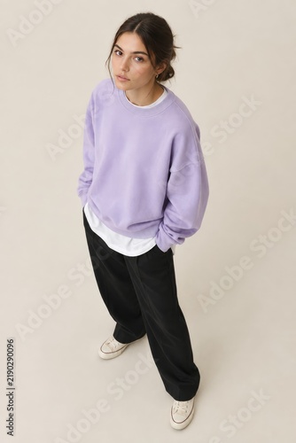 Young woman in casual chic outfit standing confidently with hands in pockets, light lavender oversized sweater layered over white tee, black wide-leg trousers, white sneakers, neutral studio