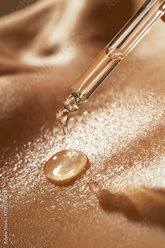 Macro close-up of a glass dropper applying golden skincare serum onto glowing skin under warm, soft light.