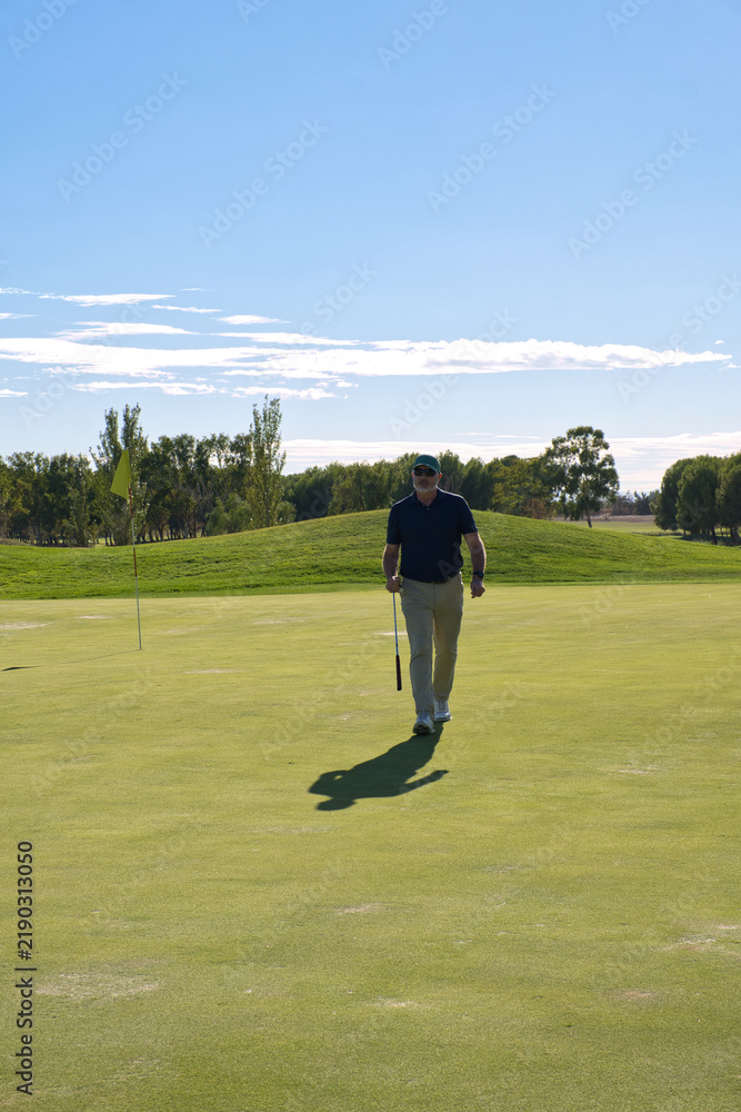 Obraz premium Senior golfer walking confidently across the green with his club in hand, flag and hole behind him - satisfaction and active retirement lifestyle.