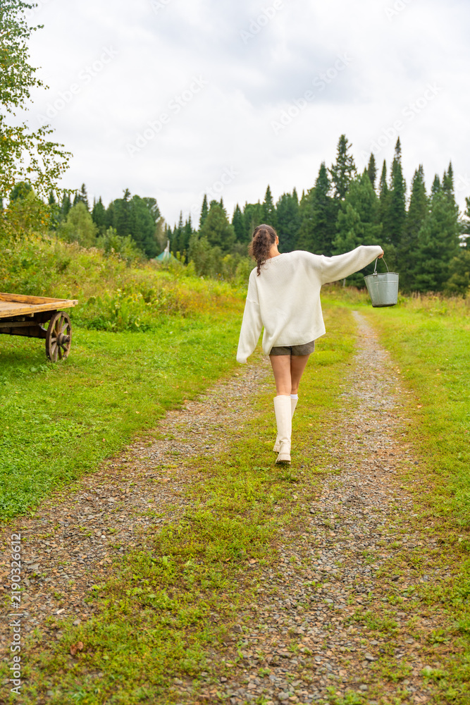 Obraz premium Young woman in white sweater and shorts walks along a gravel path carrying a bucket, with a wooden cart and trees in the background