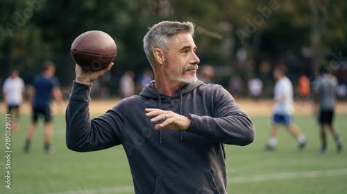 Middle-aged man in hoodie playing american football  throwing ball in city park. Gray hair  athletic man  recreational sports practice  active outdoor lifestyle  blurred players in background.
