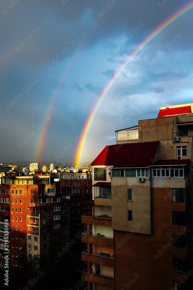Obraz premium Vibrant double rainbow arching over residential cityscape during sunset with dramatic storm clouds. Urban landscape featuring bright sunlight illumination on building facades. Concept of hope, optimis