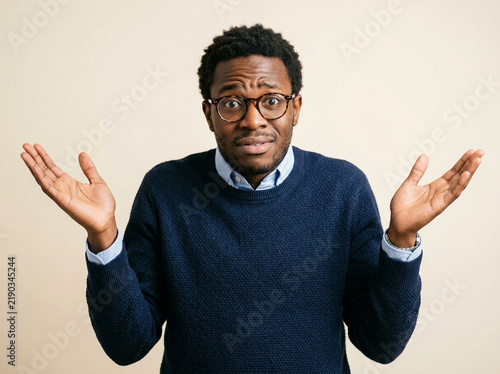 Confused black man shrugging shoulders with uncertainty against plain background
