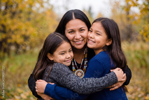 Affectionate Native American mother embracing two happy young daughters outdoors in autumn park
