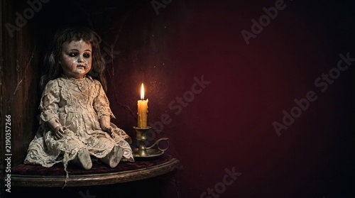 Creepy vintage porcelain doll sitting next to a lit candle in a dark haunted room with copy space