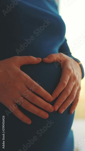 A woman gently holds her pregnant belly, forming a heart shape with her hands. The soft light creates a warm atmosphere around her and her belly.