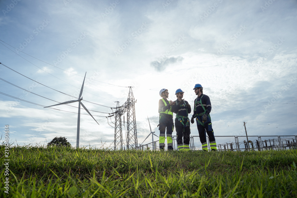 Obraz premium A group of workers stand outside near power lines and wind turbines on a sunny day. The team wears safety gear and talks together while working on a renewable energy project in the field.