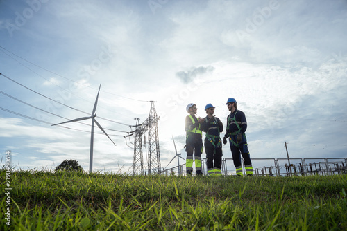 A group of workers stand outside near power lines and wind turbines on a sunny day. The team wears safety gear and talks together while working on a renewable energy project in the field.