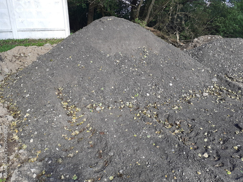 A pile of crushed stone on the ground near a concrete fence. Raw material for construction work. A dark mound of earth mixed with stones and leaves. Horizontal photo.