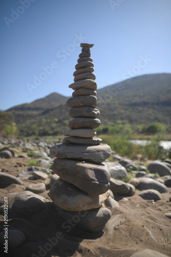 Canvas Print A tall, vertical stack of balanced river stones forming a cairn sits on a sandy shore, with a soft-focus background of a riverbed, green trees, and rolling mountains under a blue sky