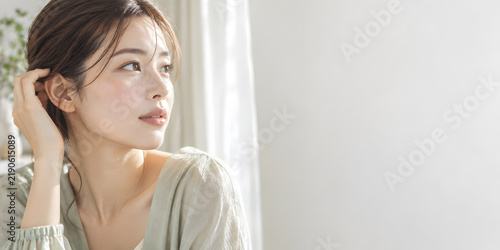美容とスキンケアを印象づける透明感のある肌 窓辺で髪を耳にかける女性と柔らかな朝の光を生かしたコピースペース