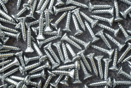 Pile of galvanized metal screws scattered on dark background. Close-up of silver self-tapping fasteners with flat countersunk heads.