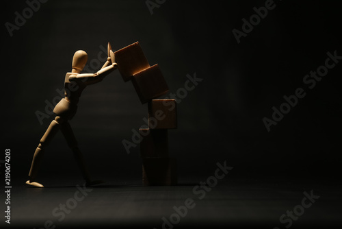 Bild auf Leinwand Wooden mannequin struggling to push or hold a falling stack of wooden cubes on dark background, concept of crisis management, heavy workload, risk, or preventing failure