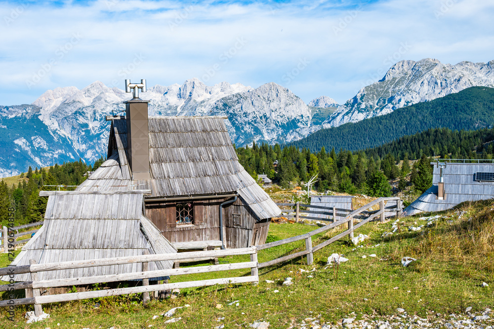 Obraz premium Alpine wooden cottage on Velika Planina set against the dramatic backdrop of the Kamnik-Savinja Alps under a clear blue sky, Slovenia