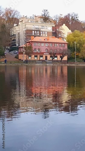 Vertical shot of European style residential building among colorful autumnal foliage trees on shoreline of city pond with calm mirror water surface at cloudy autumn day. Peaceful fall season landscape