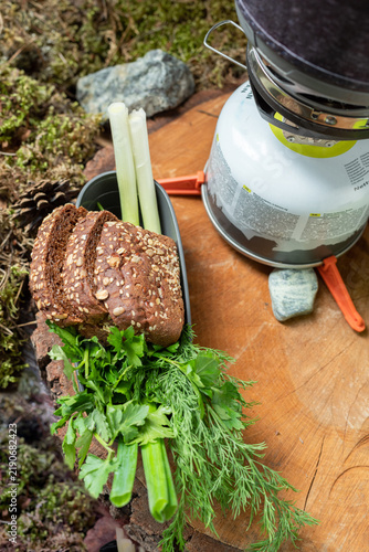 Vertical view of an outdoor camping food setup on a wooden log slice with a gas stove system, dishes with fresh green herbs, and bread.