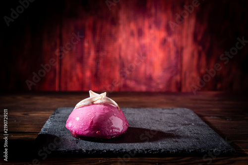 A round pink mousse pastry served on a dark black slate board against a vibrant red background. Elegant gourmet dessert presentation, French.