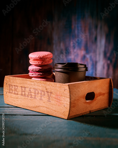 Stacked colorful macarons and coffee in a black craft paper cup inside a wooden box with Ukrainian text Be Happy. Dark mood low key lighting.