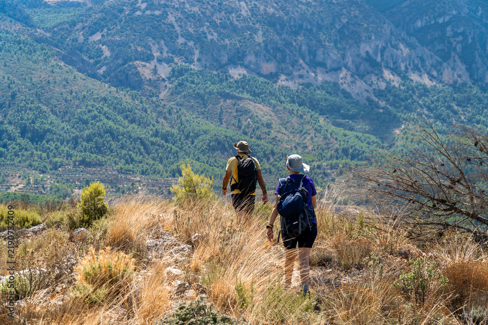 Obraz premium Two hikers walking on arid landscape with mountains on a summer day with sun and clouds