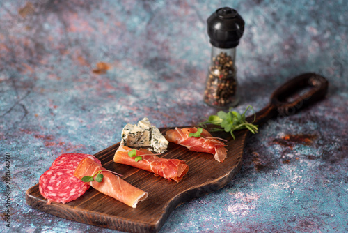 Charcuterie board with rolled jamon, sliced salami, and creamy Brie cheese on a wooden platter. Pepper mill in the soft background.
