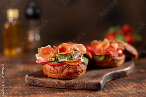 Gourmet sandwich with creamy Brie cheese, rolled jamon slices, and fresh salad greens on a wooden board. More tapas and an olive oil bottle.