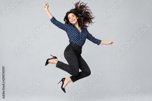 Optimistic young woman in a blue blouse and black trousers jumping happily on a grey background.