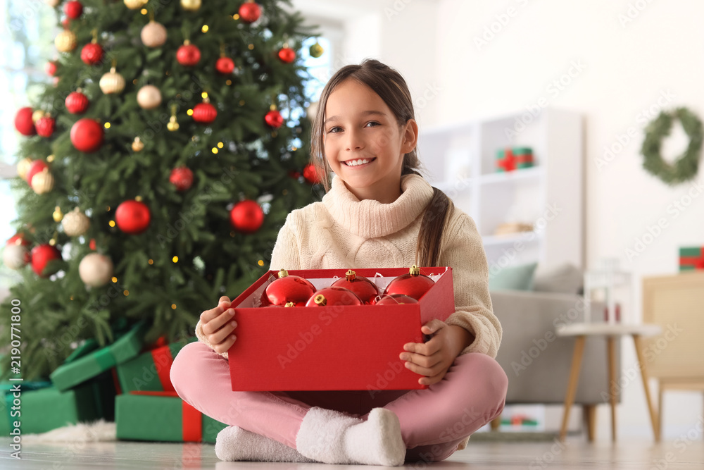 Obraz premium Smiling young girl holding a box of red christmas ornaments in front of a decorated christmas tree at home, She smiles at the camera, with a decorated christmas tree visible in the background