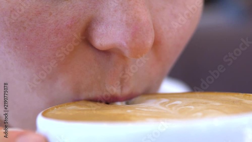 Young adult woman tasting latte, gently sipping foam topped coffee with concentrated expression in close up, coffee tasting in cozy cafe, holding white ceramic cup near mouth with fingertips visible.