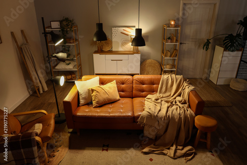 Interior of living room with shelving units, commode, armchair, glowing lamps...